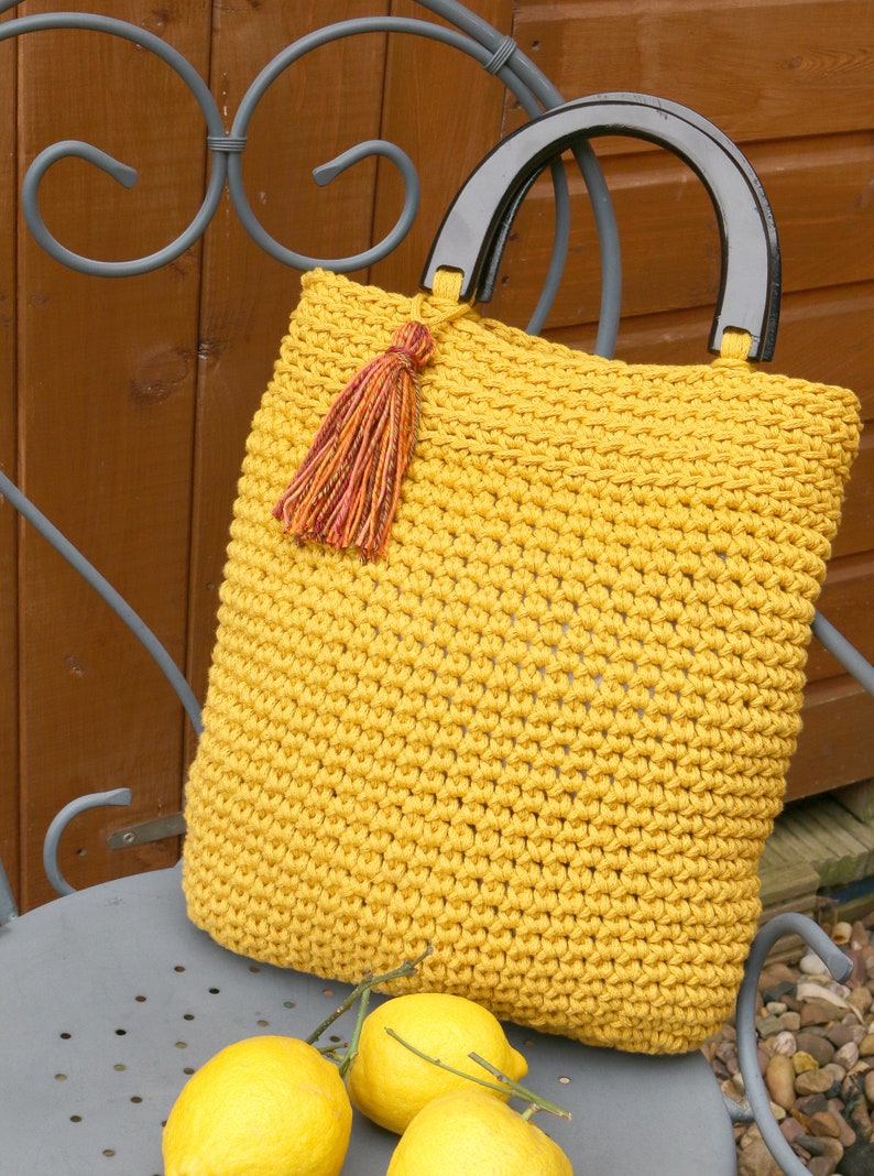 May include: A mustard yellow crocheted handbag featuring a textured stitch pattern, a dark brown semi-circular handle, and a decorative multi-colored tassel attached to the top. The bag has a square shape and is crafted from thick yarn.