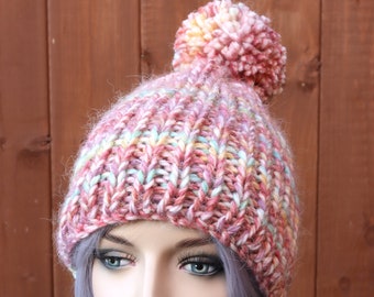 May include: A chunky knit beanie featuring a variegated yarn pattern in shades of pink, yellow, and light blue. The hat is constructed with a thick, textured rib stitch and is topped with a large, matching pom-pom made from the same multicolored yarn.
