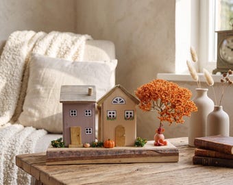 May include: A decorative wooden display featuring two small house models, one mauve and one beige, mounted on a natural wood slab base. The scene includes a miniature orange tree, tiny pumpkins, and green foliage accents arranged in front of the houses.