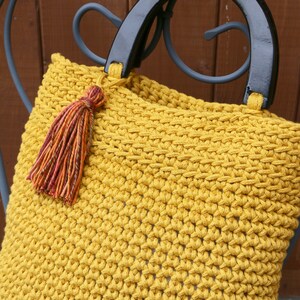 May include: A mustard yellow crocheted handbag featuring a textured stitch pattern, two glossy black semi-circular handles, and a decorative multi-colored tassel in shades of orange, red, and brown attached to one of the handle bases.