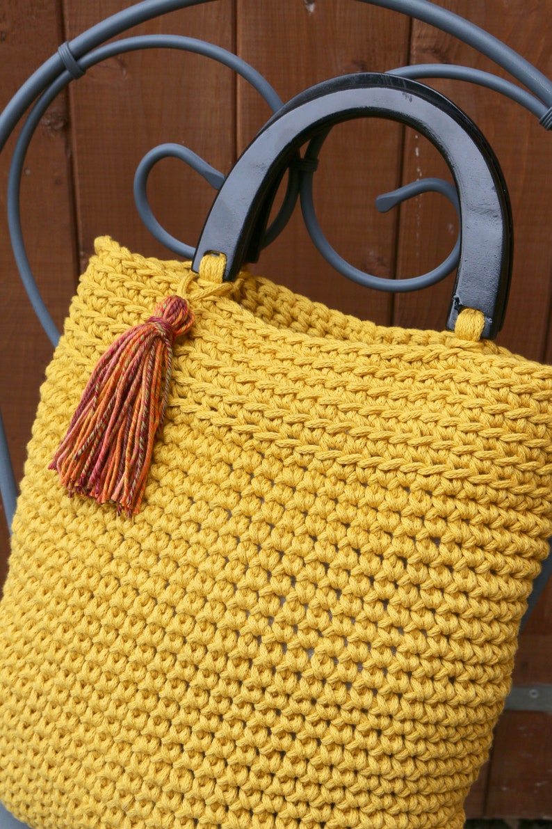 May include: A mustard yellow crocheted handbag featuring a textured stitch pattern, two glossy black semi-circular handles, and a decorative multi-colored tassel in shades of orange, red, and brown attached to one of the handle bases.