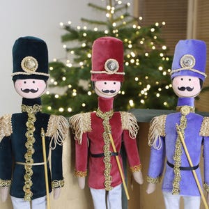 May include: Three toy soldier ornaments in a row, each with a different coloured uniform: dark green, red, and blue. They have matching moustaches, hats, and gold trim. Each soldier holds a gold staff and wears white trousers.