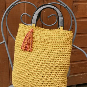 May include: A mustard yellow crocheted tote bag featuring a textured stitch pattern, two semi-circular black wooden handles, and a decorative multi-colored tassel attached to one side of the handle base, creating a distinct contrast in color.