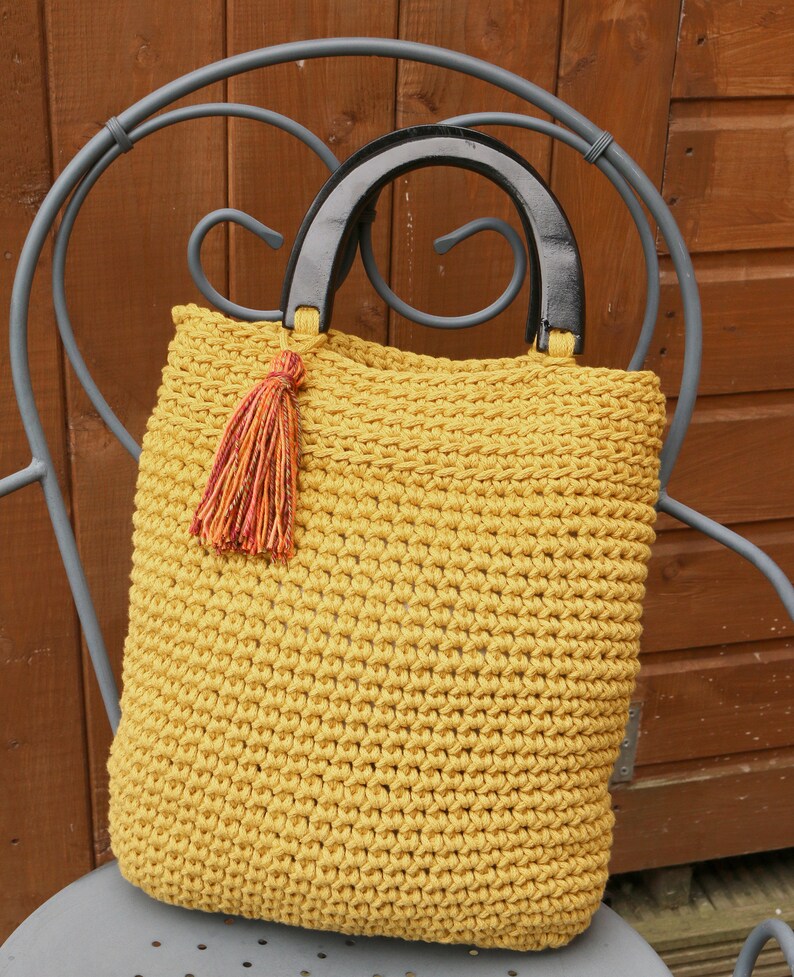 May include: A mustard yellow crocheted tote bag featuring a textured stitch pattern, two semi-circular black wooden handles, and a decorative multi-colored tassel attached to one side of the handle base, creating a distinct contrast in color.