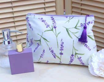May include: A rectangular fabric pouch featuring a white background with a repeating lavender floral pattern in shades of purple and green. The pouch has a purple zipper closure and a matching purple tassel attached to the zipper pull.