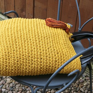 May include: A mustard yellow crocheted handbag featuring a dense, textured stitch pattern. The bag includes two sturdy, glossy black semi-circular handles and is accented with a small, multi-colored tassel in shades of orange, red, and brown.