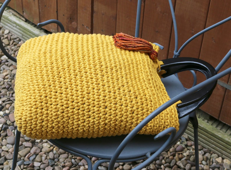 May include: A mustard yellow crocheted handbag featuring a dense, textured stitch pattern. The bag includes two sturdy, glossy black semi-circular handles and is accented with a small, multi-colored tassel in shades of orange, red, and brown.