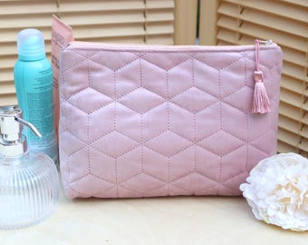 May include: A dusty rose-colored rectangular cosmetic pouch featuring a quilted geometric pattern of stitched diamonds and hexagons. The pouch includes a matching pink fabric tassel attached to the silver-toned metal zipper pull on the top right.