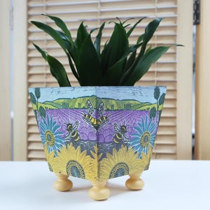 May include: A square planter with a vibrant, nature-inspired design. The planter showcases a lavender field, sunflowers, and bees. It stands on three small, rounded wooden feet and contains a green plant. The design is printed on the planter.