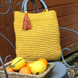 May include: A mustard yellow crocheted handbag featuring two black semi-circular handles and a decorative multi-colored tassel attached to one side. The bag has a textured, knotted stitch pattern and a rectangular shape with rounded corners.