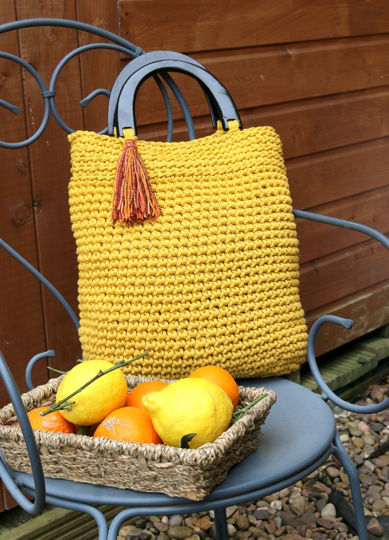 May include: A mustard yellow crocheted handbag featuring two black semi-circular handles and a decorative multi-colored tassel attached to one side. The bag has a textured, knotted stitch pattern and a rectangular shape with rounded corners.