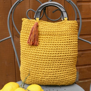 May include: A mustard yellow crocheted tote bag featuring a textured stitch pattern, two dark brown semi-circular wooden handles, and a decorative multi-colored yarn tassel attached to one side of the handle base for added detail.