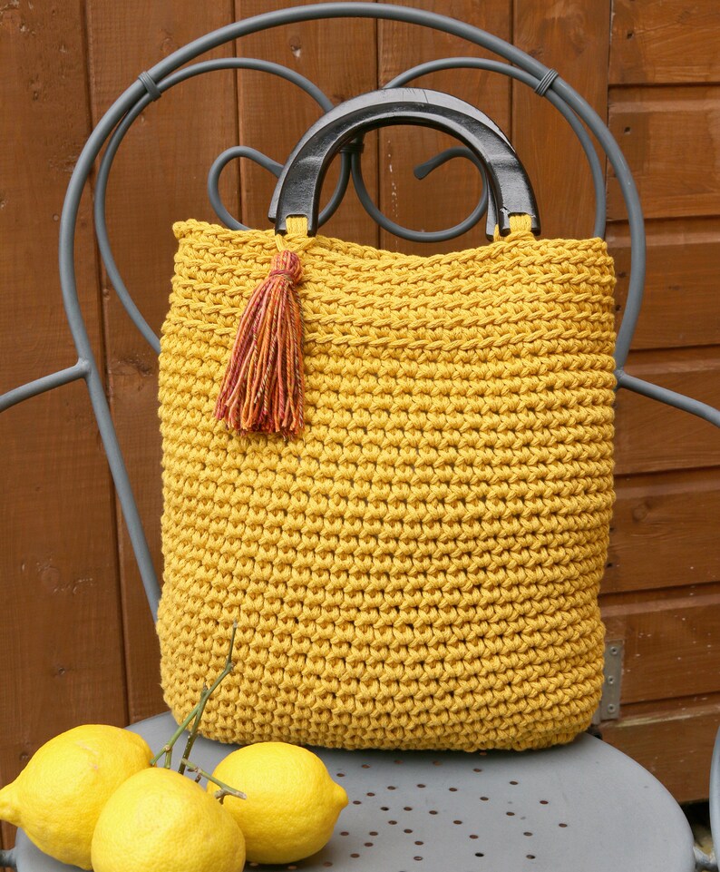 May include: A mustard yellow crocheted tote bag featuring a textured stitch pattern, two dark brown semi-circular wooden handles, and a decorative multi-colored yarn tassel attached to one side of the handle base for added detail.
