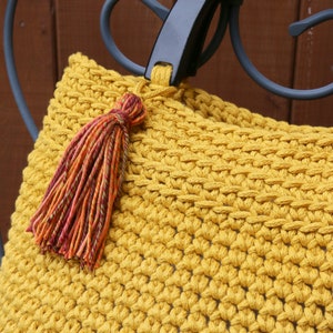 May include: A mustard yellow crocheted handbag featuring a dense, textured stitch pattern. The bag includes a smooth, black, semi-circular rigid handle and a decorative tassel made of multi-colored yarn in shades of orange, red, and green.