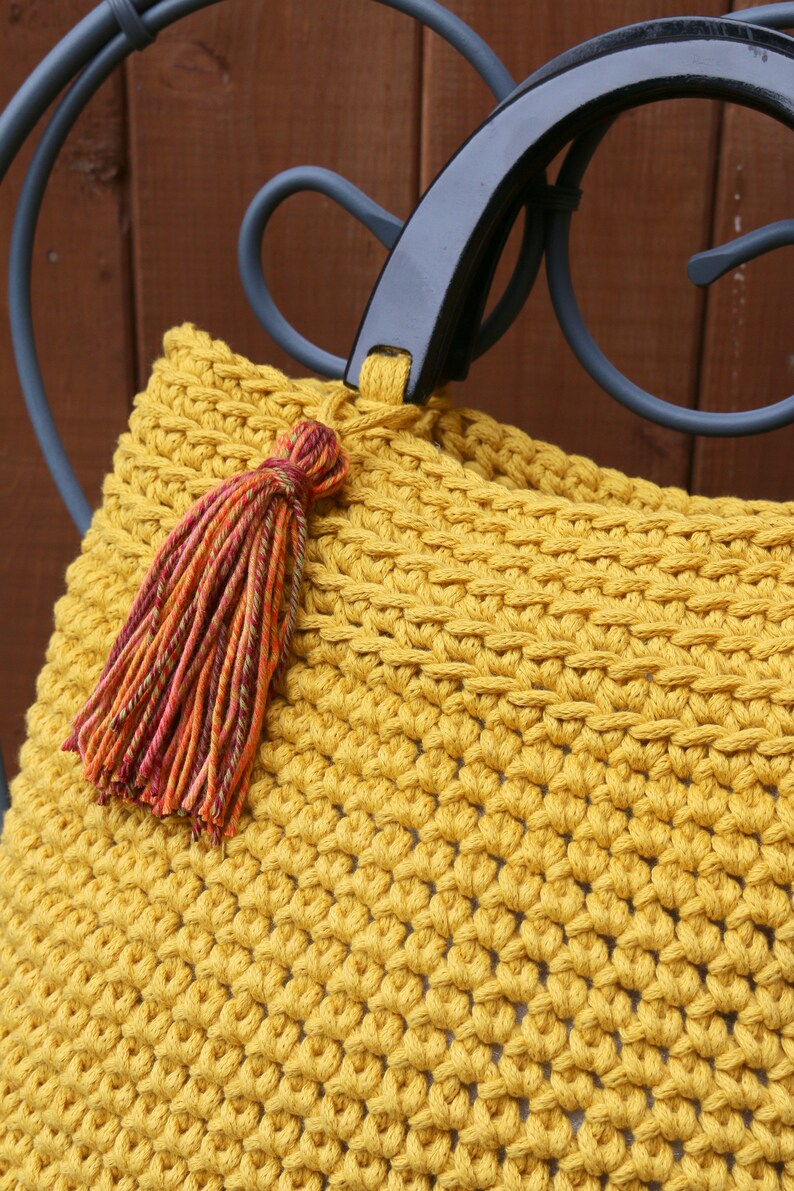 May include: A mustard yellow crocheted handbag featuring a dense, textured stitch pattern. The bag includes a smooth, black, semi-circular rigid handle and a decorative tassel made of multi-colored yarn in shades of orange, red, and green.
