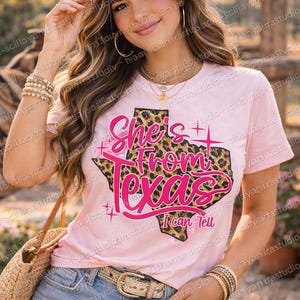 May include: A pink t-shirt with the text "She's From Texas I can Tell" in a pink font, with a leopard print Texas outline. The model is wearing a straw hat and denim shorts.