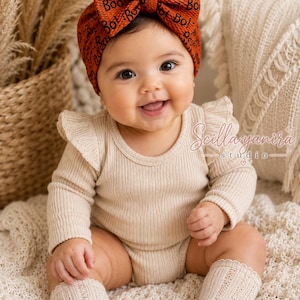 May include: A long-sleeved beige ribbed bodysuit with ruffled shoulders, paired with matching beige knit socks and a bright orange headwrap featuring a large bow and a repeating black "Boo!" text pattern across the textured fabric.