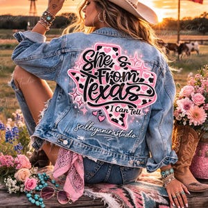 K&ouml;nnte beinhalten: Hellblau gewaschene Jeansjacke mit einem Grafikaufdruck auf dem R&uuml;cken mit der Aufschrift "She's From Texas I Can Tell" in Schwarz und Pink. Die Jacke wird mit Jeansshorts und einem Cowboyhut getragen. Der Hintergrund zeigt einen Sonnenuntergang, eine Windm&uuml;hle und eine Texas-Flagge.
