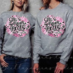 K&ouml;nnte beinhalten: Zwei graue Sweatshirts mit dem Text "She's From Texas I Can Tell" in einem rosa-schwarzen Design in Form des Bundesstaates Texas. Das Design enth&auml;lt Herzen und Sterne. Die Sweatshirts werden von zwei Personen getragen.