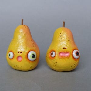 Könnte beinhalten: Zwei skurrile, handgefertigte birnenförmige Figuren. Jede Birne ist gelb mit orangefarbenen Akzenten bemalt und hat ein überraschtes Gesicht mit großen Kulleraugen und einem kleinen Mund. Die Birnen haben einen Stiel und stehen vor einem grauen Hintergrund.