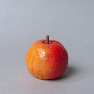 Puede incluir: Una peque&ntilde;a manzana decorativa redonda, con un degradado de colores de amarillo-naranja a rojo. Tiene una superficie texturizada y un peque&ntilde;o tallo marr&oacute;n. Es probable que la manzana sea un art&iacute;culo artesanal o decorativo.