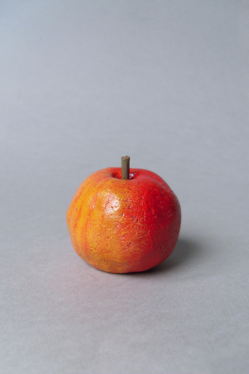 Puede incluir: Una peque&ntilde;a manzana decorativa redonda, con un degradado de colores de amarillo-naranja a rojo. Tiene una superficie texturizada y un peque&ntilde;o tallo marr&oacute;n. Es probable que la manzana sea un art&iacute;culo artesanal o decorativo.