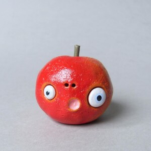 Puede incluir: Una escultura caprichosa en forma de manzana roja con una expresi&oacute;n de sorpresa. La escultura presenta grandes ojos saltones con pupilas azules, una peque&ntilde;a nariz y una boca rosada. Un peque&ntilde;o tallo marr&oacute;n se encuentra en la parte superior de la manzana.