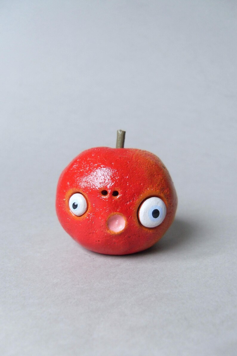 Puede incluir: Una escultura caprichosa en forma de manzana roja con una expresi&oacute;n de sorpresa. La escultura presenta grandes ojos saltones con pupilas azules, una peque&ntilde;a nariz y una boca rosada. Un peque&ntilde;o tallo marr&oacute;n se encuentra en la parte superior de la manzana.