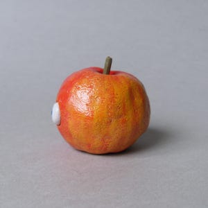 Puede incluir: Una escultura caprichosa de manzana naranja y roja con un peque&ntilde;o ojo blanco y azul adjunto. La manzana tiene una superficie texturizada y un peque&ntilde;o tallo marr&oacute;n.