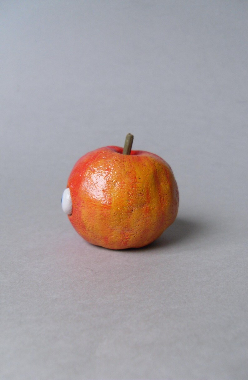 Puede incluir: Una escultura caprichosa de manzana naranja y roja con un peque&ntilde;o ojo blanco y azul adjunto. La manzana tiene una superficie texturizada y un peque&ntilde;o tallo marr&oacute;n.
