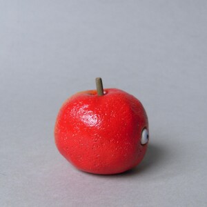 Puede incluir: Una manzana roja brillante con una superficie texturizada y un peque&ntilde;o tallo marr&oacute;n. La manzana presenta un solo ojo de dibujos animados con un dise&ntilde;o blanco y azul. Este art&iacute;culo decorativo es una versi&oacute;n l&uacute;dica de una fruta cl&aacute;sica.