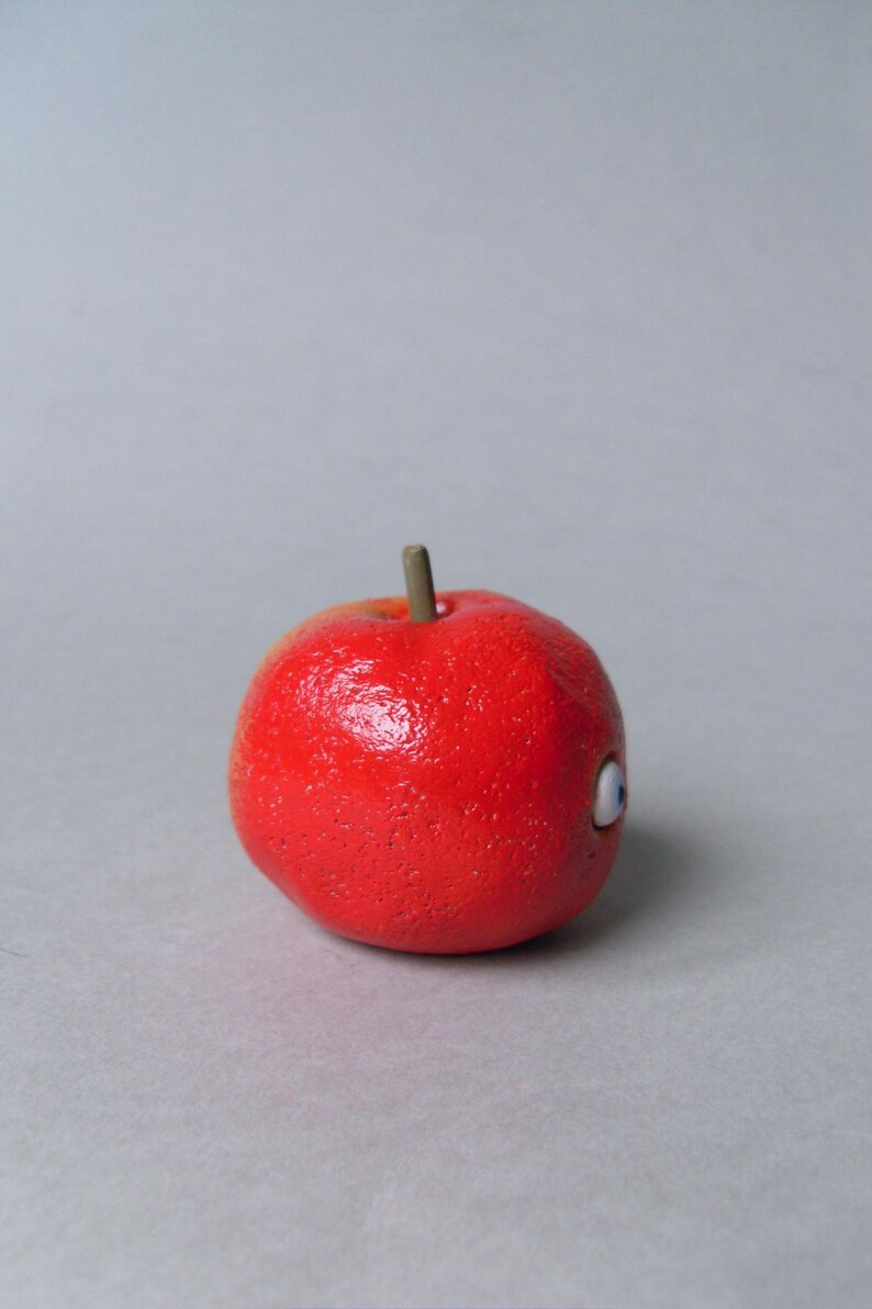 Puede incluir: Una manzana roja brillante con una superficie texturizada y un peque&ntilde;o tallo marr&oacute;n. La manzana presenta un solo ojo de dibujos animados con un dise&ntilde;o blanco y azul. Este art&iacute;culo decorativo es una versi&oacute;n l&uacute;dica de una fruta cl&aacute;sica.