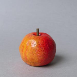 Puede incluir: Una peque&ntilde;a manzana artificial redonda con una superficie texturizada. La manzana pasa de un rojo vibrante en la parte superior a un naranja c&aacute;lido en la parte inferior. Un peque&ntilde;o tallo marr&oacute;n sobresale de la parte superior. El fondo es gris claro s&oacute;lido.