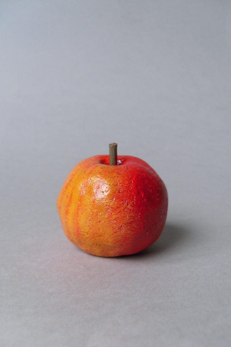Puede incluir: Una peque&ntilde;a manzana artificial redonda con una superficie texturizada. La manzana pasa de un rojo vibrante en la parte superior a un naranja c&aacute;lido en la parte inferior. Un peque&ntilde;o tallo marr&oacute;n sobresale de la parte superior. El fondo es gris claro s&oacute;lido.