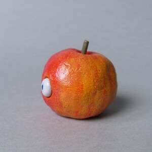 Puede incluir: Una escultura caprichosa, pintada a mano, de una manzana con un solo ojo de dibujos animados. La manzana es principalmente roja y naranja, con un tallo verde. El ojo es blanco con un iris azul, a&ntilde;adiendo un toque juguet&oacute;n a la fruta.