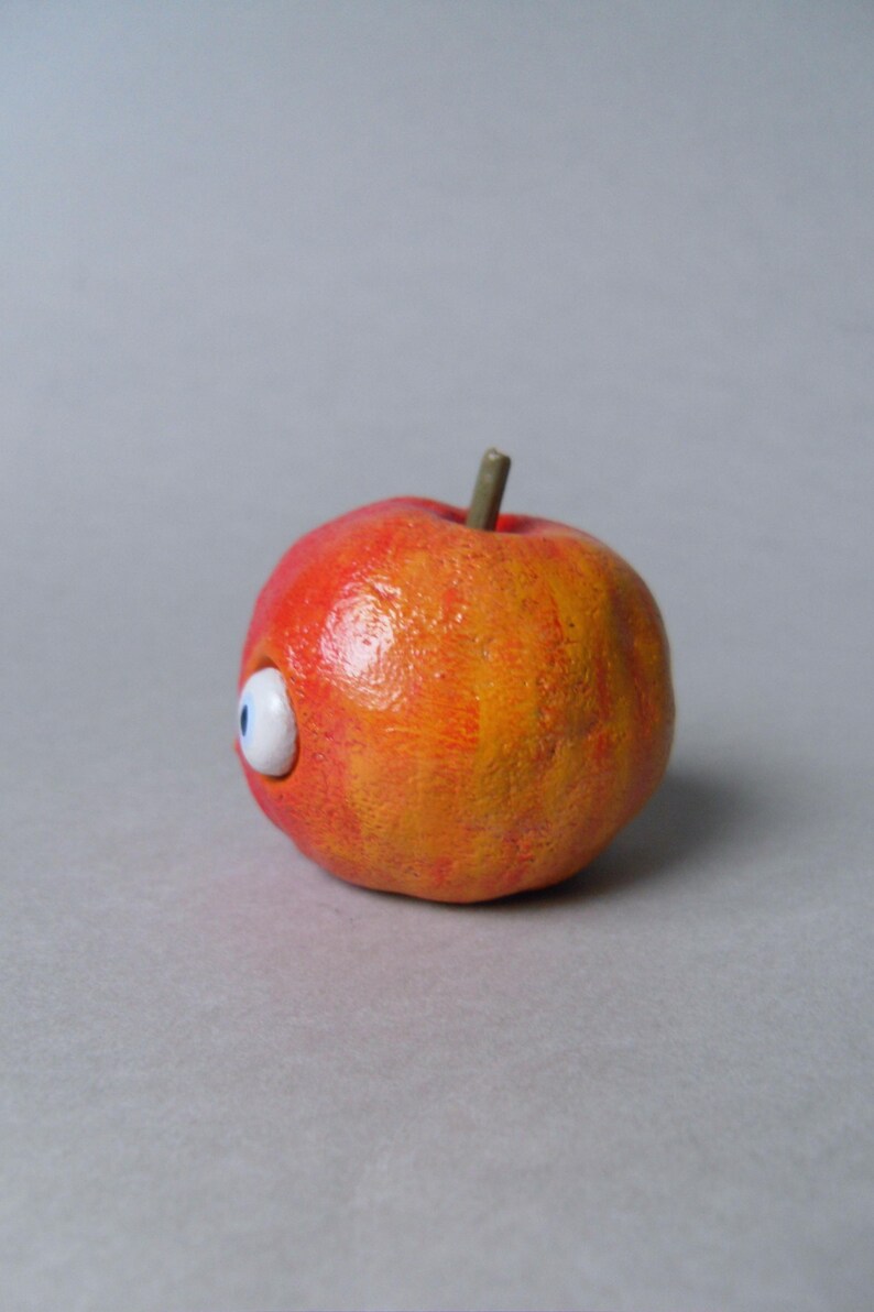 Puede incluir: Una escultura caprichosa, pintada a mano, de una manzana con un solo ojo de dibujos animados. La manzana es principalmente roja y naranja, con un tallo verde. El ojo es blanco con un iris azul, a&ntilde;adiendo un toque juguet&oacute;n a la fruta.