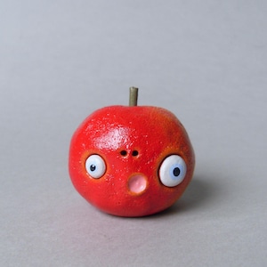 Puede incluir: Una escultura redonda y roja caprichosa que representa una manzana con una cara. Presenta grandes ojos blancos y azules, una peque&ntilde;a nariz y una boca rosada. La escultura tiene una superficie texturizada y un peque&ntilde;o tallo marr&oacute;n.