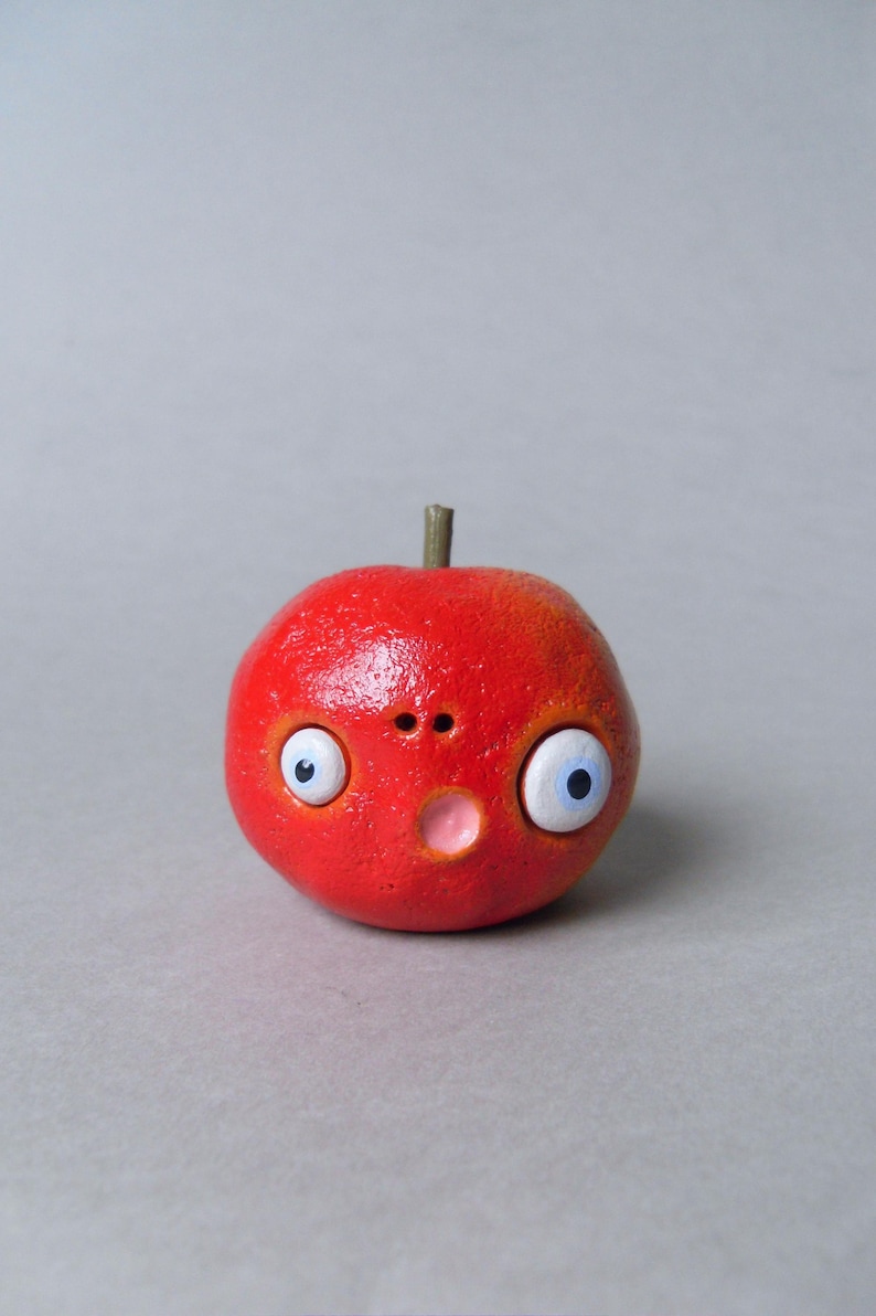 Puede incluir: Una escultura redonda y roja caprichosa que representa una manzana con una cara. Presenta grandes ojos blancos y azules, una peque&ntilde;a nariz y una boca rosada. La escultura tiene una superficie texturizada y un peque&ntilde;o tallo marr&oacute;n.