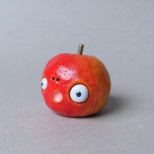 Puede incluir: Una escultura artesanal caprichosa de una manzana roja con una expresi&oacute;n de sorpresa. La manzana presenta grandes ojos de dibujos animados, una peque&ntilde;a nariz y una boca abierta. El tallo es de color marr&oacute;n oscuro.