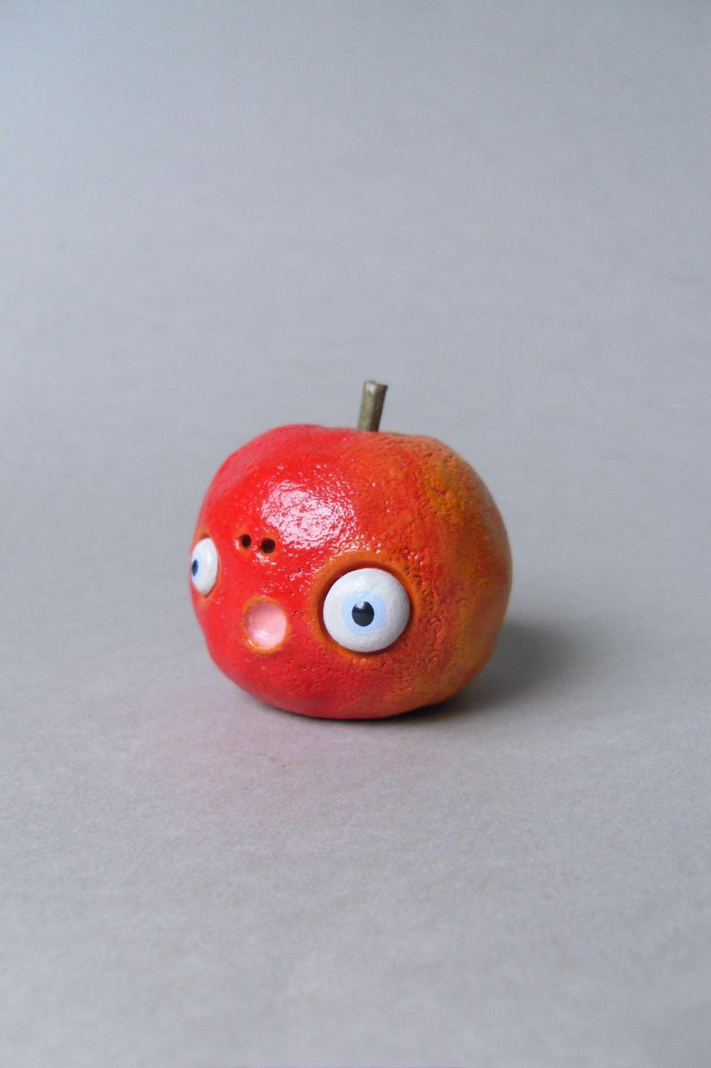 Puede incluir: Una escultura artesanal caprichosa de una manzana roja con una expresi&oacute;n de sorpresa. La manzana presenta grandes ojos de dibujos animados, una peque&ntilde;a nariz y una boca abierta. El tallo es de color marr&oacute;n oscuro.