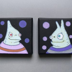 Puede incluir: Dos pequeñas pinturas de lienzo negro con personajes caprichosos y de dibujos animados. Los personajes son de color azul claro con ojos grandes y llevan camisas a rayas. El fondo es negro con lunares morados y rosas.