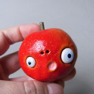 Puede incluir: Una escultura redonda roja caprichosa con rasgos de dibujos animados. Tiene ojos grandes con bordes blancos e iris azules, una nariz peque&ntilde;a y una boca rosada. Un peque&ntilde;o tallo marr&oacute;n est&aacute; en la parte superior. La escultura se sostiene en una mano.