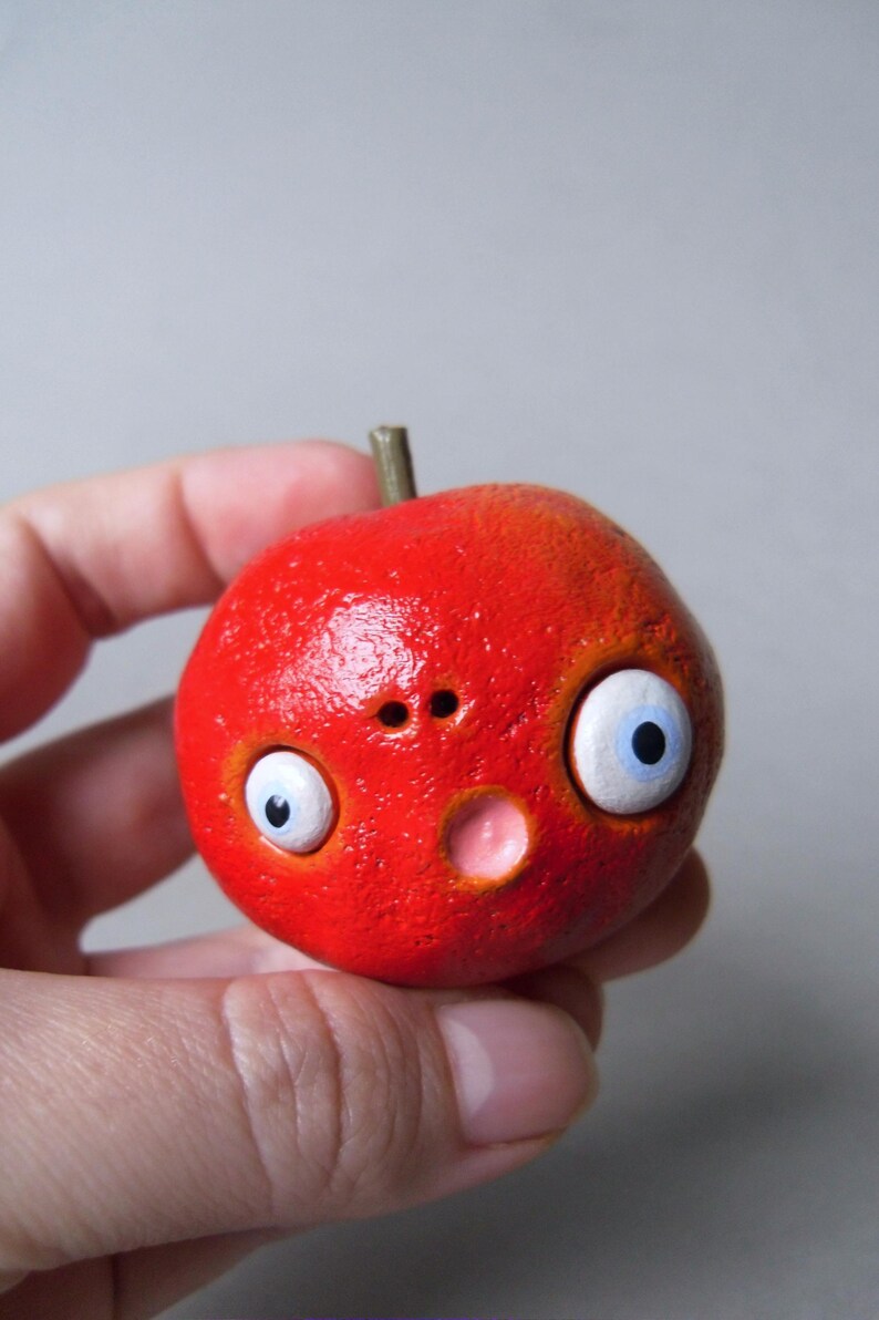 Puede incluir: Una escultura redonda roja caprichosa con rasgos de dibujos animados. Tiene ojos grandes con bordes blancos e iris azules, una nariz peque&ntilde;a y una boca rosada. Un peque&ntilde;o tallo marr&oacute;n est&aacute; en la parte superior. La escultura se sostiene en una mano.