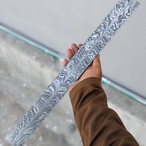 Puede incluir: Una larga pieza rectangular de acero con un patrón arremolinado único en tonos grises y blancos. El acero tiene una apariencia de acero de Damasco, con un diseño ondulado y veteado. El artículo se sostiene en ángulo, mostrando el intrincado patrón. Mide unos 30 cm.