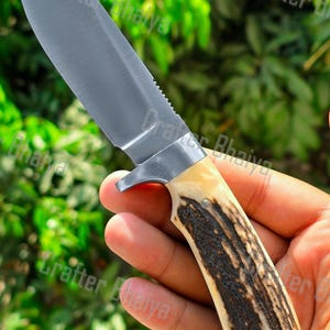 Puede incluir: Un cuchillo de caza con una hoja de acero pulido y un lomo dentado. El mango está hecho de material estampado marrón claro y marrón oscuro. El texto "Crafter Bhaiya" es visible en la hoja.