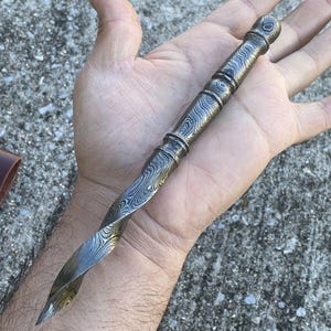 May include: A Damascus steel dagger with a twisted blade and patterned handle. The dagger has a pointed tip and a decorative, textured design. The handle features a series of rings and a rounded end.