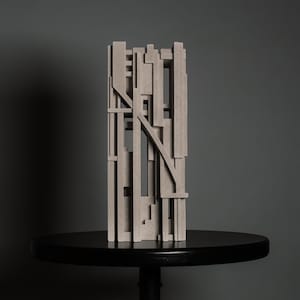 May include: A tall, rectangular sculpture made of light grey, textured material. The object features a complex arrangement of vertical and horizontal bars, with a single diagonal beam crossing the centre, creating an open, grid-like structure.