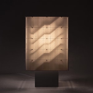 May include: A rectangular table lamp featuring a grid of square panels with diagonal lines, illuminated from within to create a warm glow. The lamp is supported by a solid, dark, rectangular base and emits light through the translucent panels.