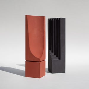 Peut inclure: Deux sculptures abstraites. L'une est une pièce haute de couleur terre cuite avec un sommet incurvé et une base carrée. L'autre est une sculpture rectangulaire noire avec un motif d'escalier. Les deux sont sur fond blanc.