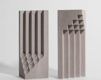 Op de afbeelding: Twee rechthoekige, lichtgrijze, gespikkelde stenen sculpturen. Het linkerstuk heeft verticale groeven die eindigen in halfronde inkepingen. Het rechterstuk toont verticale groeven met een getrapt, geometrisch uitgesneden patroon.